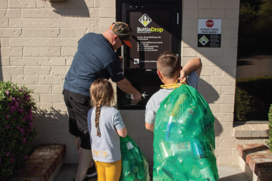 One adult and two kids putting Green Bags into a BottleDrop Redemption Center drop door
