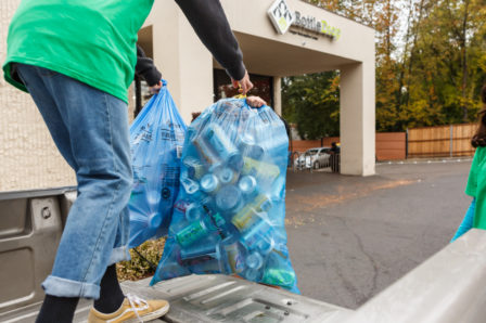 A person standing in a truck bed handing two full BottleDrop Give Blue Bags to a teammate who is going to drop off the bags at the Redemption Center drop door.