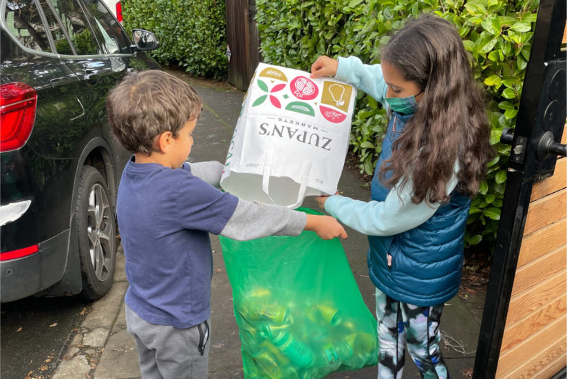 On a sunny day, 5 year old and 9 year old siblings are working together to collect bottles and cans and pour them from a grocery bag into a BottleDrop Green Bag.