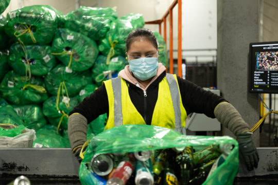 BottleDrop employee opening a Green Bag to process for a customer. A rack of two over 12 bags are in the background awaiting to be processed.